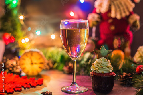 Новогодние декорации. На столе, украшенном новогодними гирляндами и Санта Клаусом, стоят деревянные часы и бокал шампанского с пирожнным