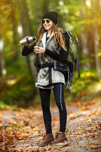 Cheerful brunette tourist girl wears black cap and sunglasses, backpacked have break with thermos of tea, autumn tourism concept