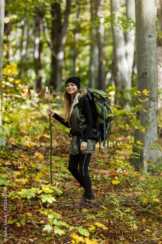 Cheerful brunette tourist girl wears black cap and backpacked have walk through forest holding stick, autumn tourism concept
