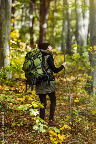 Cheerful brunette tourist girl wears black cap and backpacked have walk through forest holding stick, autumn tourism concept