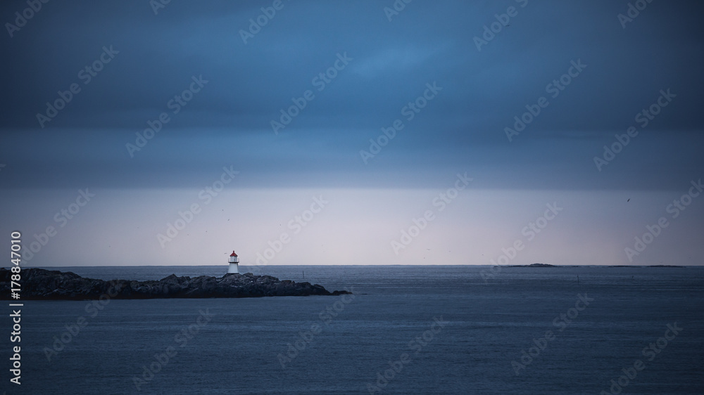 Little lighthouse on rocks surrounded by an ocean against cloudy sky
