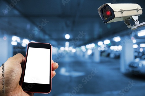hand using mobile phone with blank screen and CCTV, security indoor camera system operating in underground car parking garage area, blue color tone, surveillance security and safety technology concept