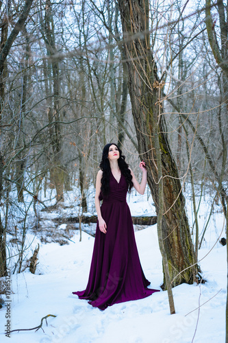 A young woman in an elegant burgundy evening dress in winter among trees and snow conceals a riddle