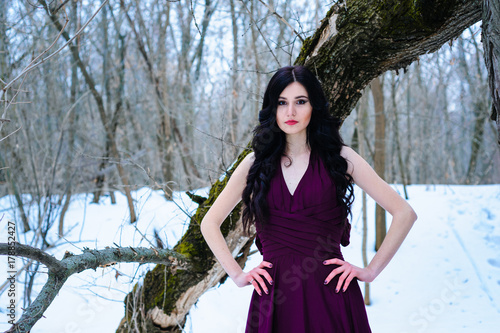 A young woman in an elegant burgundy evening dress in winter among trees and snow conceals a riddle © Alla