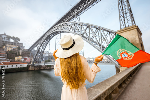Young woman traveler in sunhat standing back with portuguese flag with the famous iron bridge on the background in Porto city