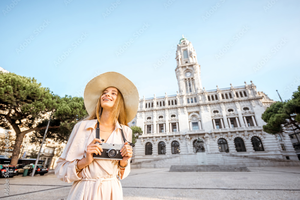 Portrait of a young woman tourist in sunhat standing with photo camera in front of the city hall building during the morning light in Porto, Portugal