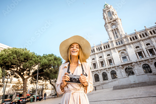 Portrait of a young woman tourist in sunhat standing with photo camera in front of the city hall building during the morning light in Porto, Portugal © rh2010