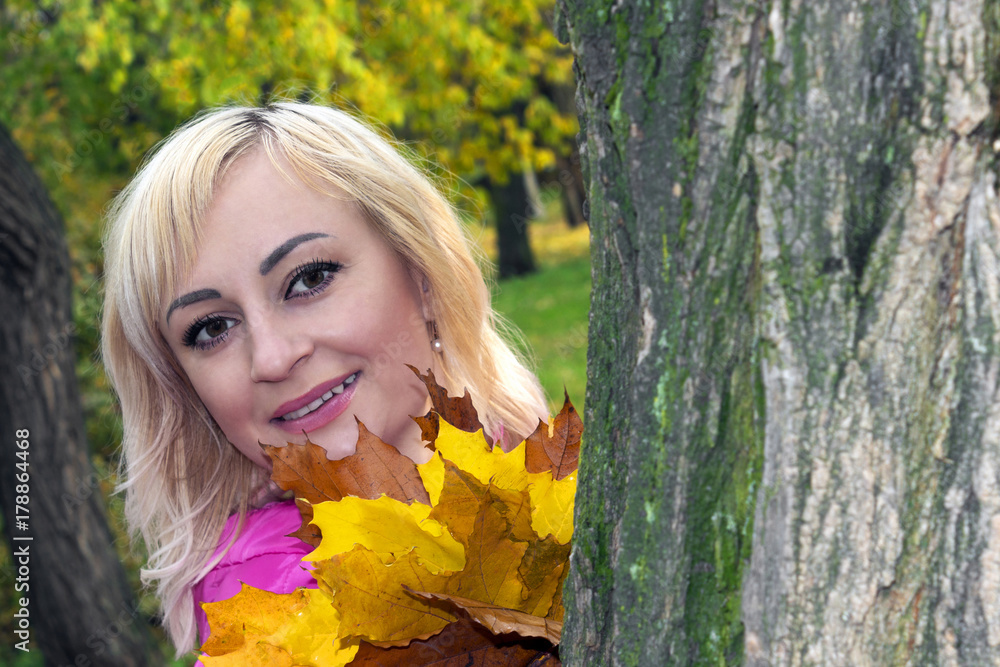 Beautiful girl, blond, peeks from behind a tree with a bouquet of maple leaves