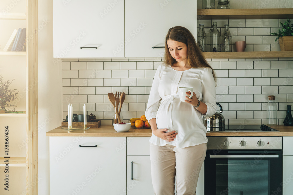A young attractive pregnant woman wearing in white shirt is standing in the kitchen, leaning on the table, drinking a drink and looking at her belly. Happy people, love, maternity, lifestyle. A young attractive pregnant woman wearing in white shirt is standing in the kitchen, leaning on the table, drinking a drink and looking at her belly. Happy people, love, maternity, lifestyle.