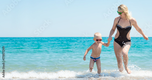 Beautiful young mother and small son holding hands running on the waves on the beach.Fun,family,friendly summer vacation. © Denis