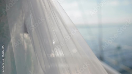 The bride stands on the yacht. A white veil develops in the wind. Closeup shot.