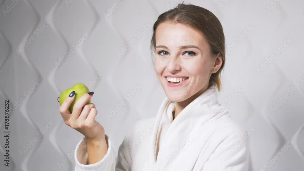 Happy woman in the white bathrobe eating an apple, indoor shot in the white background Happy woman in the white bathrobe eating an apple, indoor shot in the white background