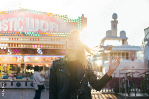 Beautiful fashion model with blonde hair, wears black leather jacket and stands in middle of carnvial, festival or fair, in front of attraction rides during summer time holidays or spring break photo