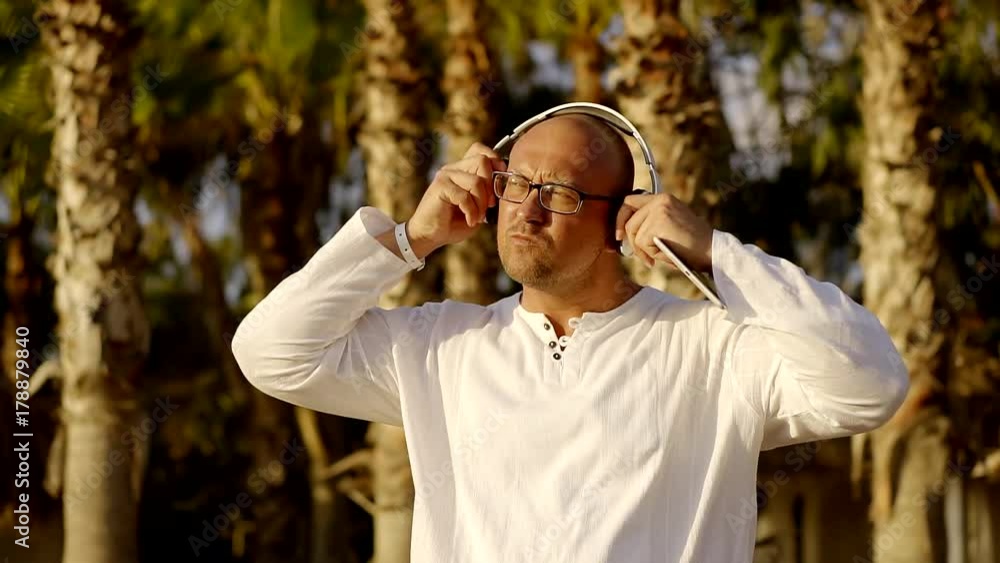 middle-aged man is putting on earphones and choosing song on display of tablet, standing near palms middle-aged man is putting on earphones and choosing song on display of tablet, standing near palms