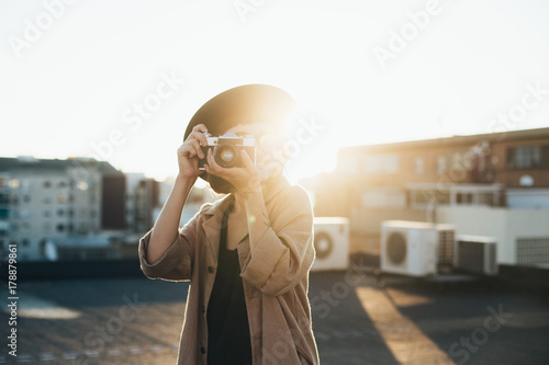 Trendy and attractive beautiful woman with analog vintage film camera makes photos on rooftop at sunset time, creates content for social media channels and applications, popular influencer photo