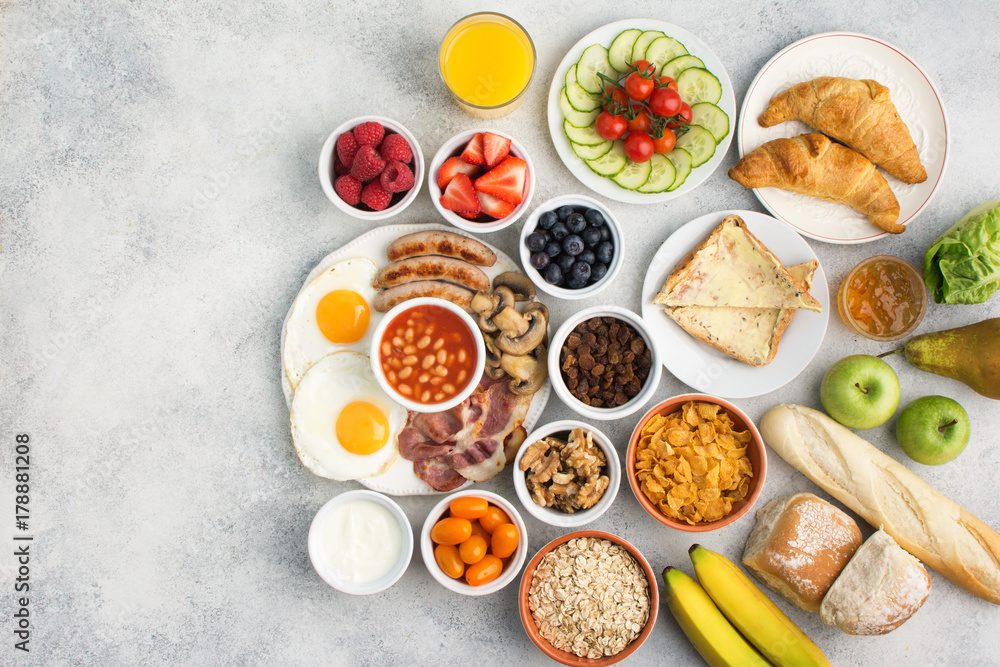 Healthy varied breakfast, fried eggs, sausages, bacon and mushrooms with selection of fruits and vegetables, breads and juice on the grey white table, top view, copy space for text, selective focus