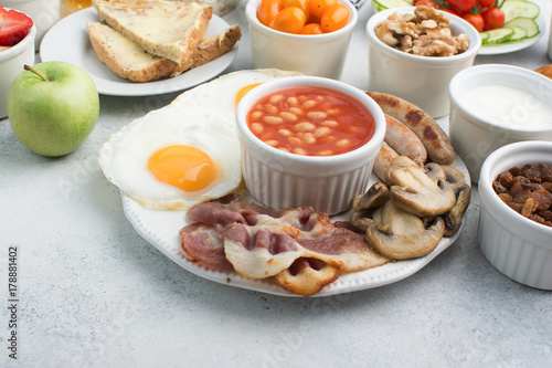 English breakfast made with fried eggs, sausages, bacon and mushrooms with selection of fruits and vegetables, breads and juice on the grey white table, copy space for text, selective focus