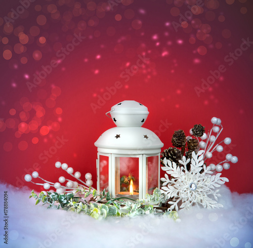 Christmas Eve lantern and decorations background. Picture of a white lantern with lighted candle decorated with pine cones on a white snow against red frost background 