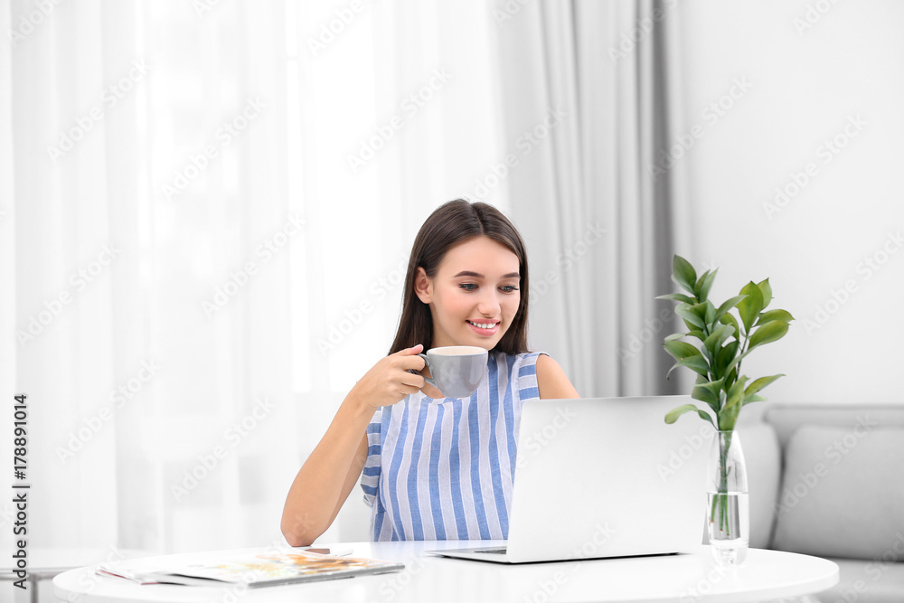 Young lady with modern laptop drinking coffee while sitting at table in room Young lady with modern laptop drinking coffee while sitting at table in room