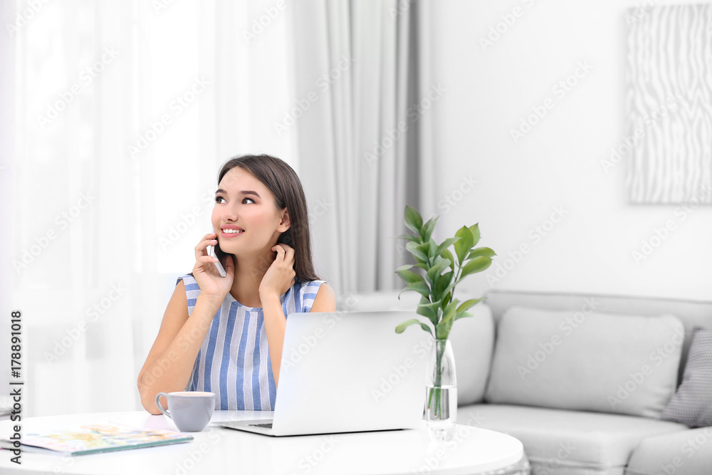 Young lady with modern laptop talking by phone while sitting at table in room Young lady with modern laptop talking by phone while sitting at table in room