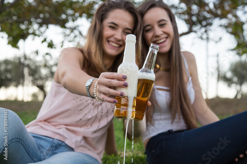 Two young laughing women posing on lawn in park and toasting with beer bottles having fun.  