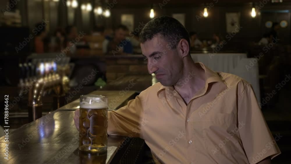 Alcoholic man drinking his sorrow alone at the bar in a pub with people on background Alcoholic man drinking his sorrow alone at the bar in a pub with people on background