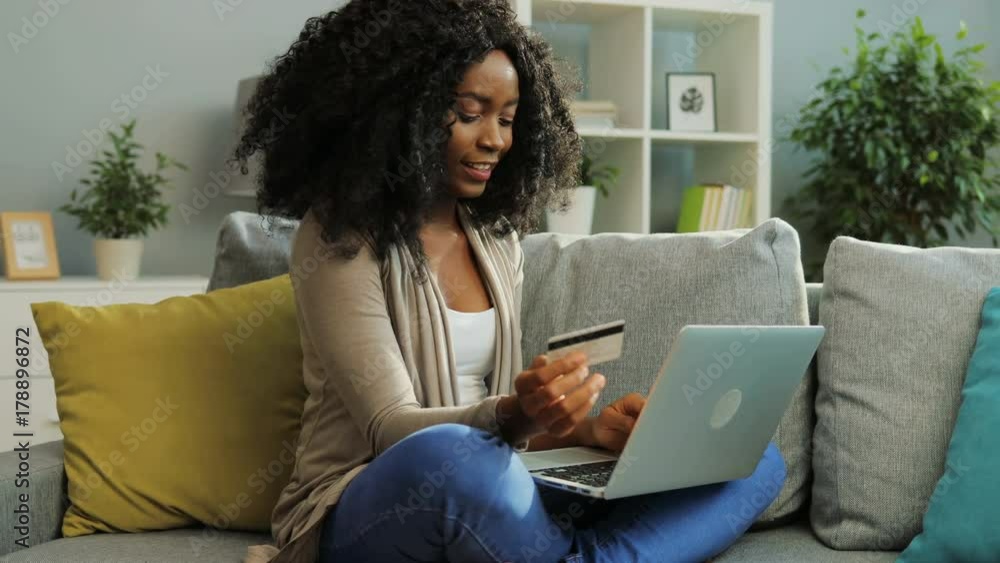 Pretty African American young woman online shopping with credit card using her laptop. Living room. Pretty African American young woman online shopping with credit card using her laptop. Living room.