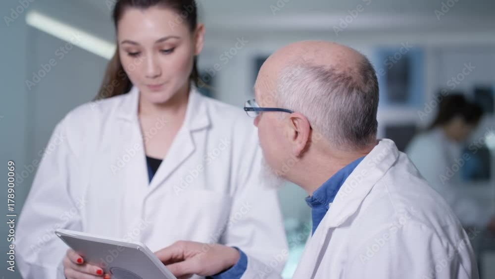 Portrait smiling senior scientist looking at tablet and discussing work with young researcher in the lab. Portrait smiling senior scientist looking at tablet and discussing work with young researcher in the lab.