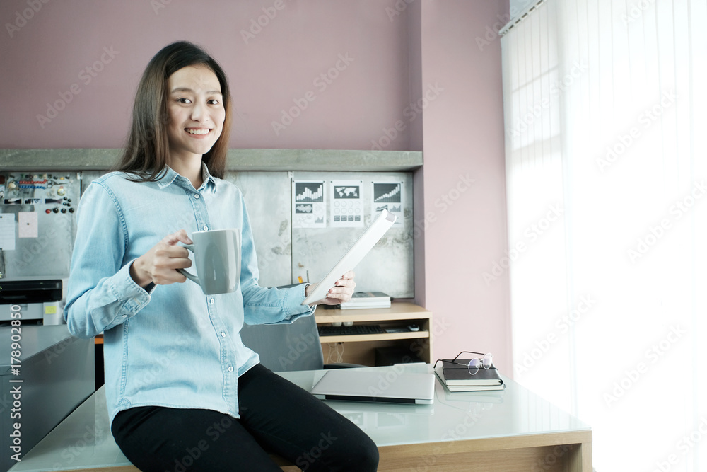 Young asian businesswoman working with tablet and holding a coffee cup with smiling face while sitting on office desk, casual office life concept Young asian businesswoman working with tablet and holding a coffee cup with smiling face while sitting on office desk, casual office life concept