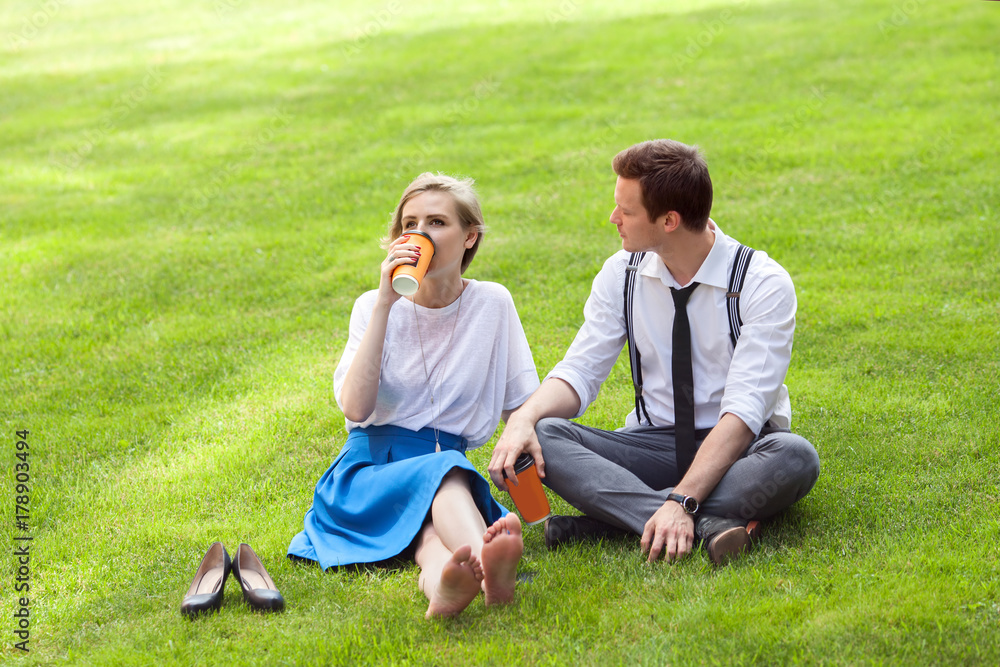 Young man and woman are drinking coffee on green meadow in city park