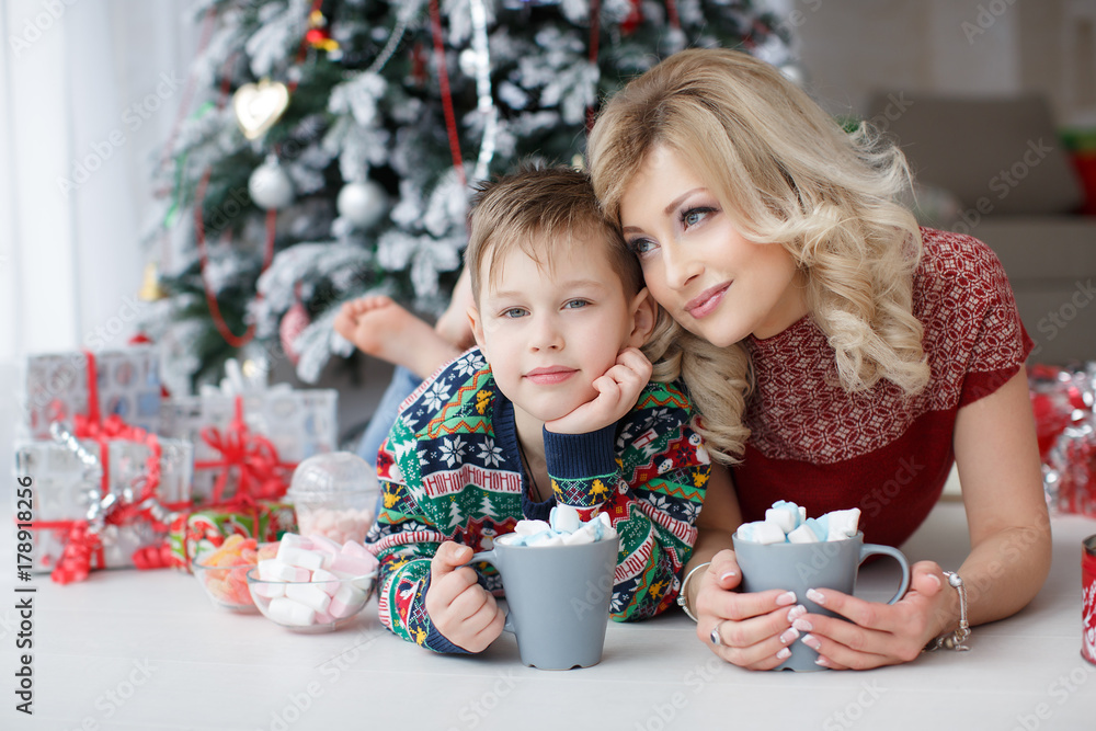 Beautiful woman and her five year old son lie side by side on a white floor in the room on the green background,elegant Christmas tree and colorful boxes with Christmas gifts,holding large grey cups Beautiful woman and her five year old son lie side by side on a white floor in the room on the green background,elegant Christmas tree and colorful boxes with Christmas gifts,holding large grey cups