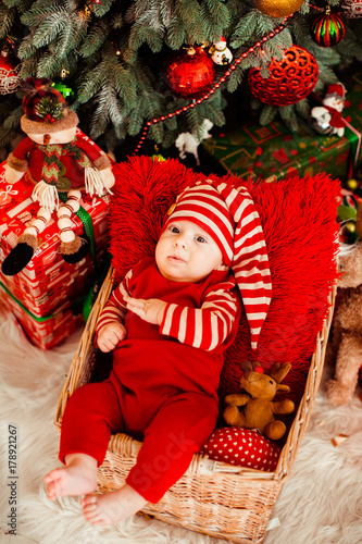 Little boy in funny red suit and long hat lies in the basket before a Christmas tree © pyrozenko13