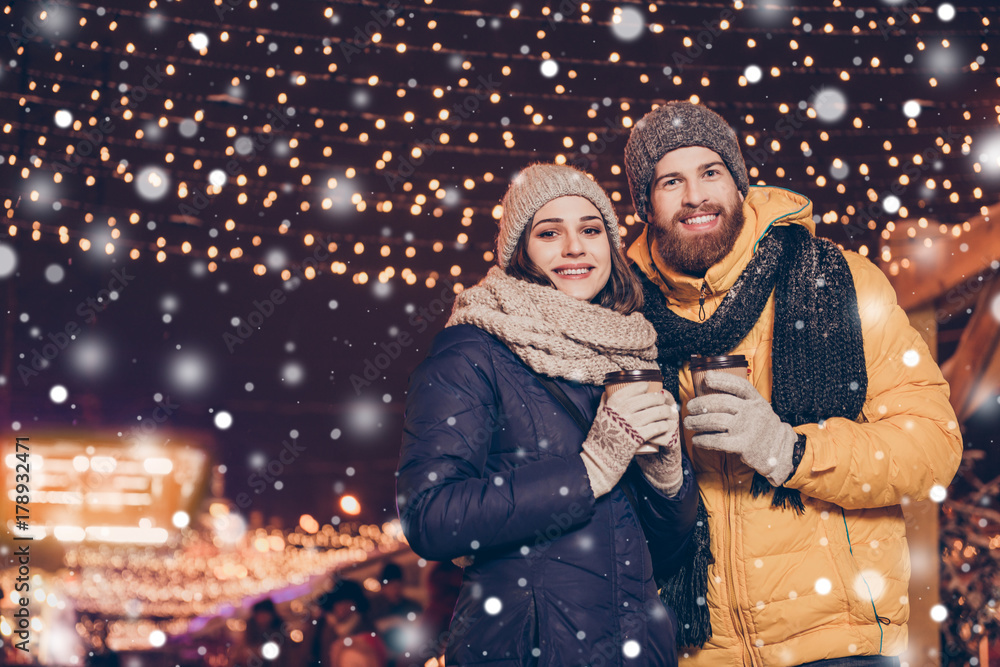 New year`s time! Low angle view of young couple in love is walking in a park of attractions outdoors, enjoying, holding cups of hot chocolate, dressed warm, christmas lights behind New year`s time! Low angle view of young couple in love is walking in a park of attractions outdoors, enjoying, holding cups of hot chocolate, dressed warm, christmas lights behind