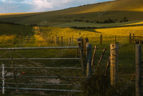 Setting sun casts long shadows on fenced hills in front of a farm gate. East Sussex  England.