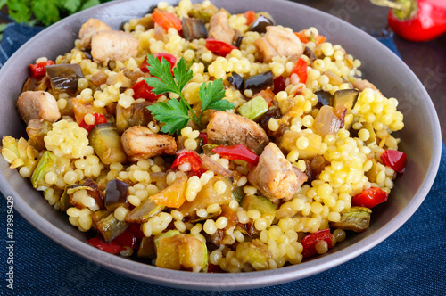 Light healthy dietary salad with couscous, vegetables (zucchini, eggplant, carrots, sweet peppers, onions), chicken pieces on a dark wooden background. Close up