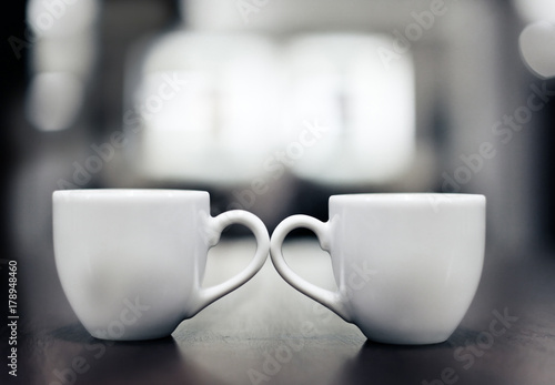 two white coffee cups standing side by side and touching their ears, in the background nice blurry