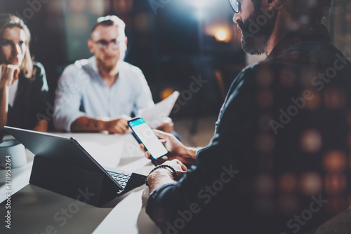 Coworkers working together in modern office.Bearded man talking with colleague and using mobile laptop while sitting at the wooden table.Horizontal.Blurred background.Flares.