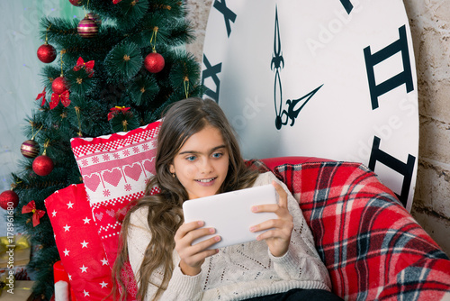 Young girl with a tablet on the couch. Winter holidays, New Year,  large clock. Interior room. girl is reading congratulations for Christmas.  girl sends online greeting cards
