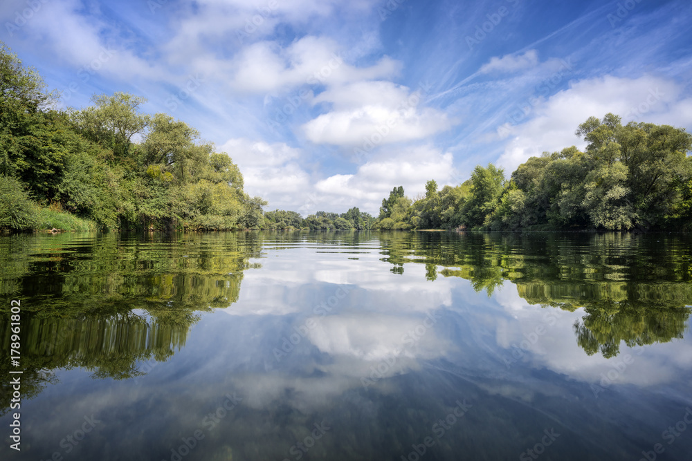 Pure nature with calm river, trees on riverbank and a dramatic blue sky which mirrors on the surface of the river