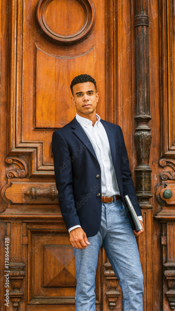 Stockfoto African American Businessman working in New York. Wearing black  blazer, white shirt, gray pants, holding laptop computer, a college student  standing by vintage office doorway on campus, looking at you
