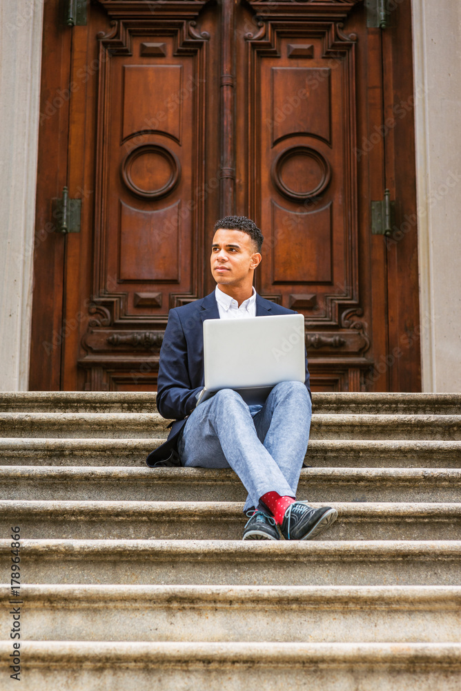 Way to Success. African American college student studying in New York, wearing blue blazer, gray pants, red socks, sneakers, crossing legs, sitting on stairs, working on laptop computer, thinking..