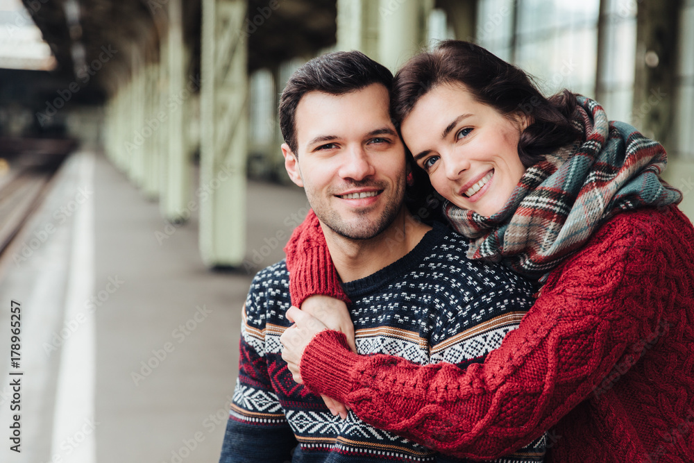 Portrait of happy young female embraces her husband, smiles happily, going to travel together, wait train on railway station, have good relationships. Romantic couple express positive emotions