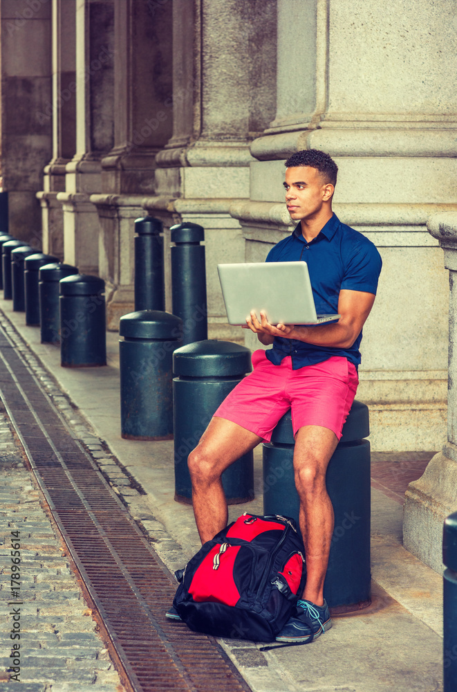 African American College Student traveling, studying in New York, wearing blue short sleeve shirt, red shorts, with backpack on ground, sitting on pillar on street, working on laptop computer..