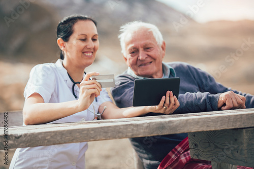 Smiling caregiver nurse and disabled senior patient using digital tablet and credit card outdoor