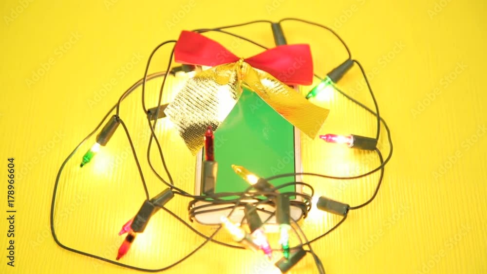 Phone as a gift for the new year. Mobile phone in a garland and bow on a yellow background with a green screen, a gift for Christmas Phone as a gift for the new year. Mobile phone in a garland and bow on a yellow background with a green screen, a gift for Christmas