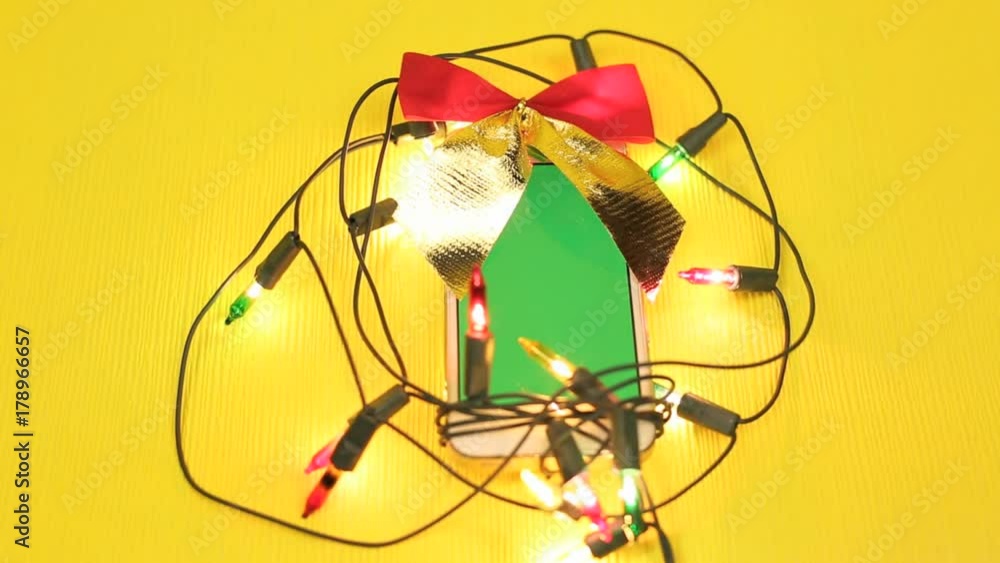 Phone as a gift for the new year. Mobile phone in a garland and bow on a yellow background with a green screen, a gift for Christmas Phone as a gift for the new year. Mobile phone in a garland and bow on a yellow background with a green screen, a gift for Christmas