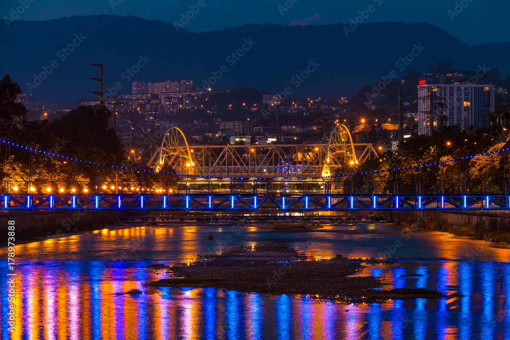Night view of illuminated Malyy Kubanskiy bridge, Sochinskiy bridge Sochi river with lights reflected in its water, Sochi, Russia
