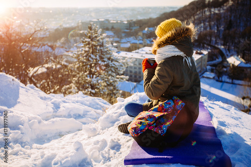 Happiness, winter holidays, tourism, travel and people concept - young woman in yellow knitting hat and red mittens drinking hot tea and sitting on the hill and looking on snowy city view background. photo