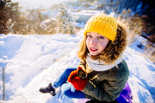 Top view portrait of happy freezing hipster girl in yellow cup and red mittens with thermo cup relaxing on the hill in snow winter sunny day. Winter holidays, tourism, travel and people concept.