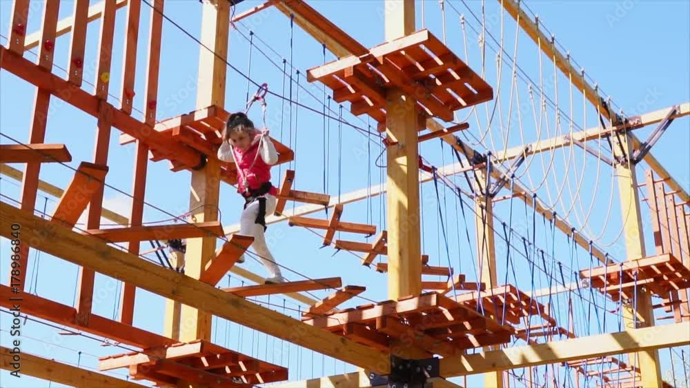 The child walks on the hanging park, the girl steps over the wooden steps and deftly copes with the obstacles, it is secured with the help of an insurance cable The child walks on the hanging park, the girl steps over the wooden steps and deftly copes with the obstacles, it is secured with the help of an insurance cable